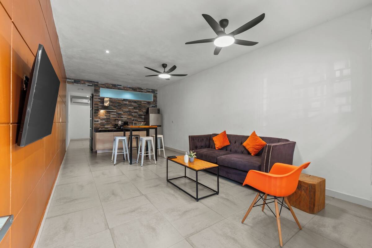 A modern living area with a sofa, orange chair, and a coffee table. There are two ceiling fans and a flat-screen TV mounted on the wall.