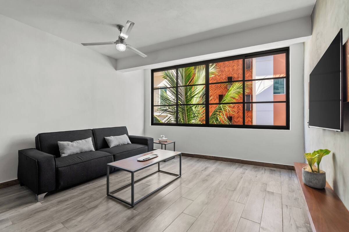 A modern living room with a black sofa, a coffee table, and a large window with a view of a brick building and greenery.