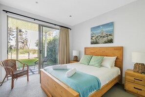 Ein Schlafzimmer mit einem großen Bett, einem Holz-Kopfteil, Blick ins Freie durch eine Schiebetür, einem Korbstuhl und einem Bild an der Wand.
