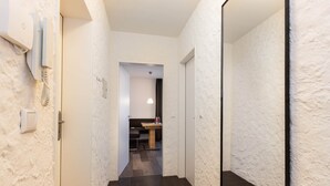 Un pasillo estrecho con paredes blancas con textura y suelo de baldosas oscuras. Una puerta a la izquierda da a un cuarto con cama y mesita de noche. Un espejo a la derecha refleja parte del pasillo.