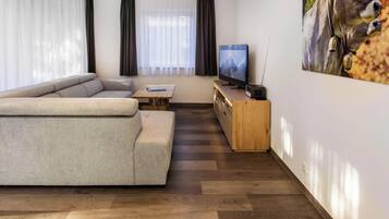 Ein modernes Wohnzimmer mit einem großen, winkelförmigen Sofa, einem Holztisch, einem Fernseher und einem kleinen Lautsprecher.