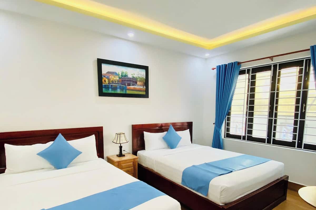 Ein Hotelzimmer mit zwei Betten, Holz-Kopfteilen, weißer Bettwäsche, blauen Akzenten, einem Nachttisch mit Lampe, einem gerahmten Bild an der Wand, einem Fenster mit Vorhängen und einem kleinen Tisch mit einem Stuhl.