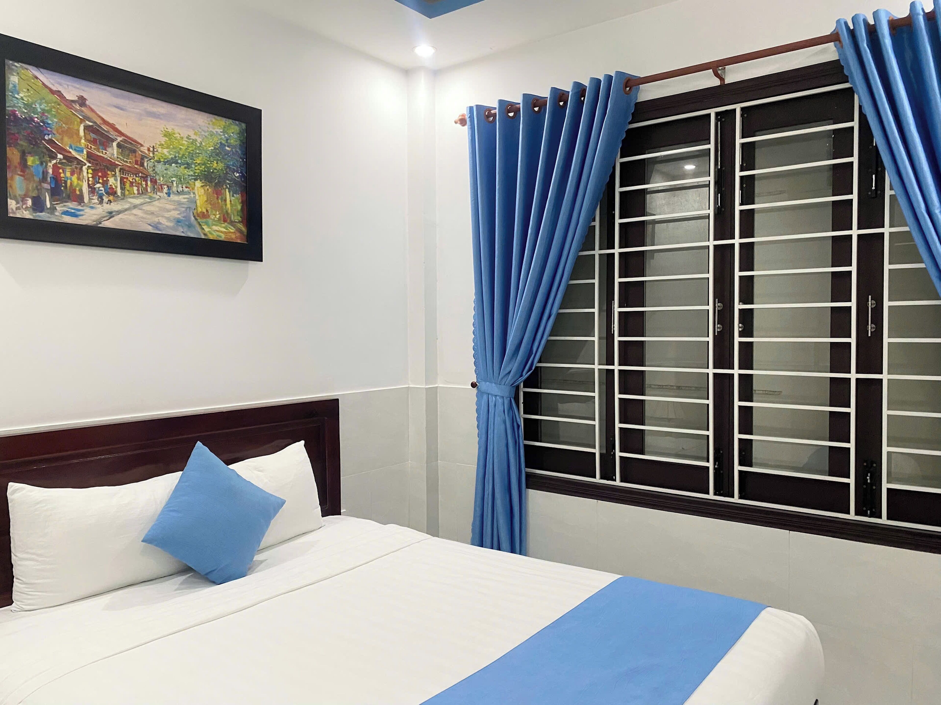 A hotel room with a bed, a window with blue curtains, and a painting on the wall.