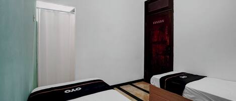 A room with two single beds, a wooden wardrobe, a door with a red curtain, and a window with white blinds.