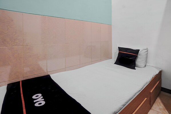 A single bed with a white mattress and two pillows, one black and one white, in a room with a tiled wall and a wooden headboard.