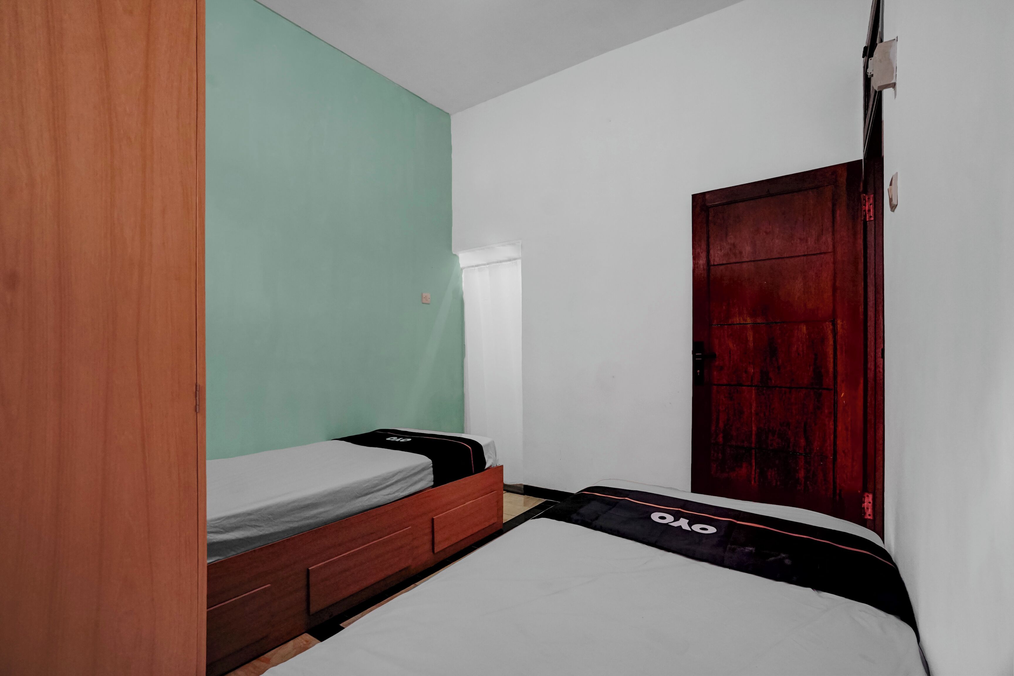 A small, single-bed room with a wooden wardrobe, a green accent wall, and a red door.