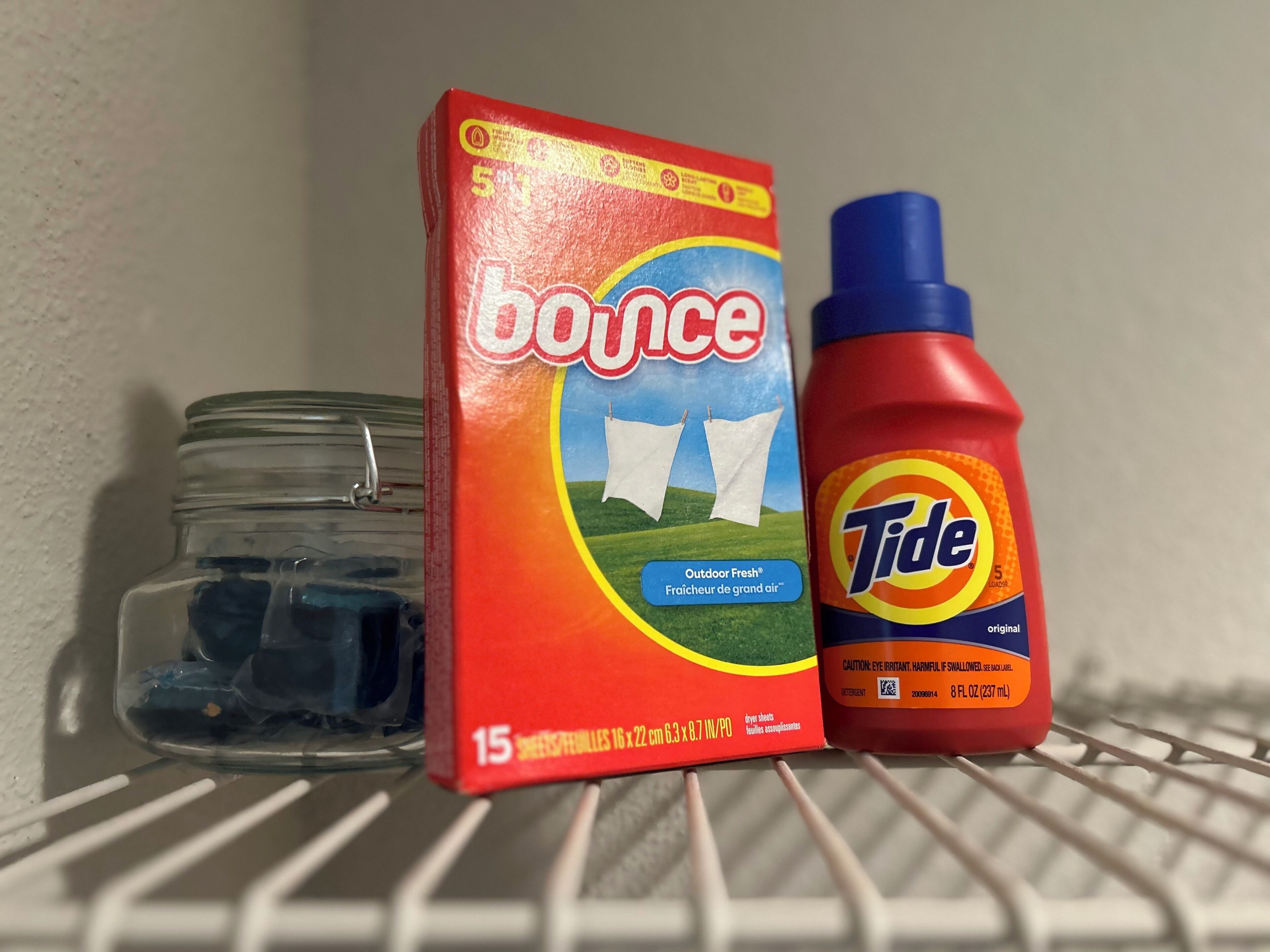 Una estantería con una caja de detergente Bounce, una botella de detergente Tide y un tarro de vidrio.