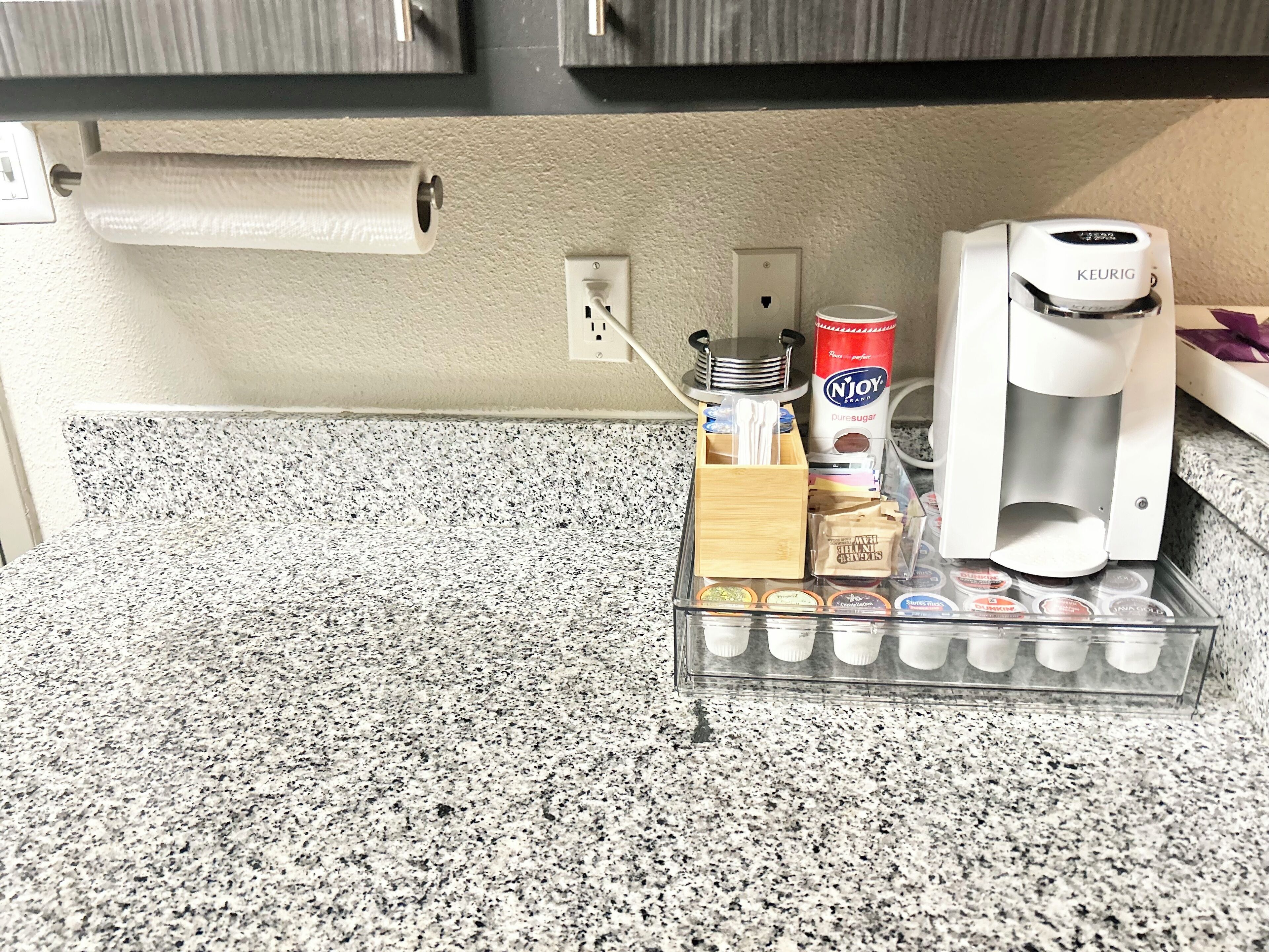 Una encimera de cocina con una cafetera, una lata de café Njoy, un tazón de azúcar y un organizador de plástico transparente con cápsulas de café.