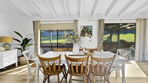 A dining area with a white table and wooden chairs, a potted plant, and a view of a green landscape through large windows. - Pet-friendly Private Retreat with Large Backyard (Surf Beach)