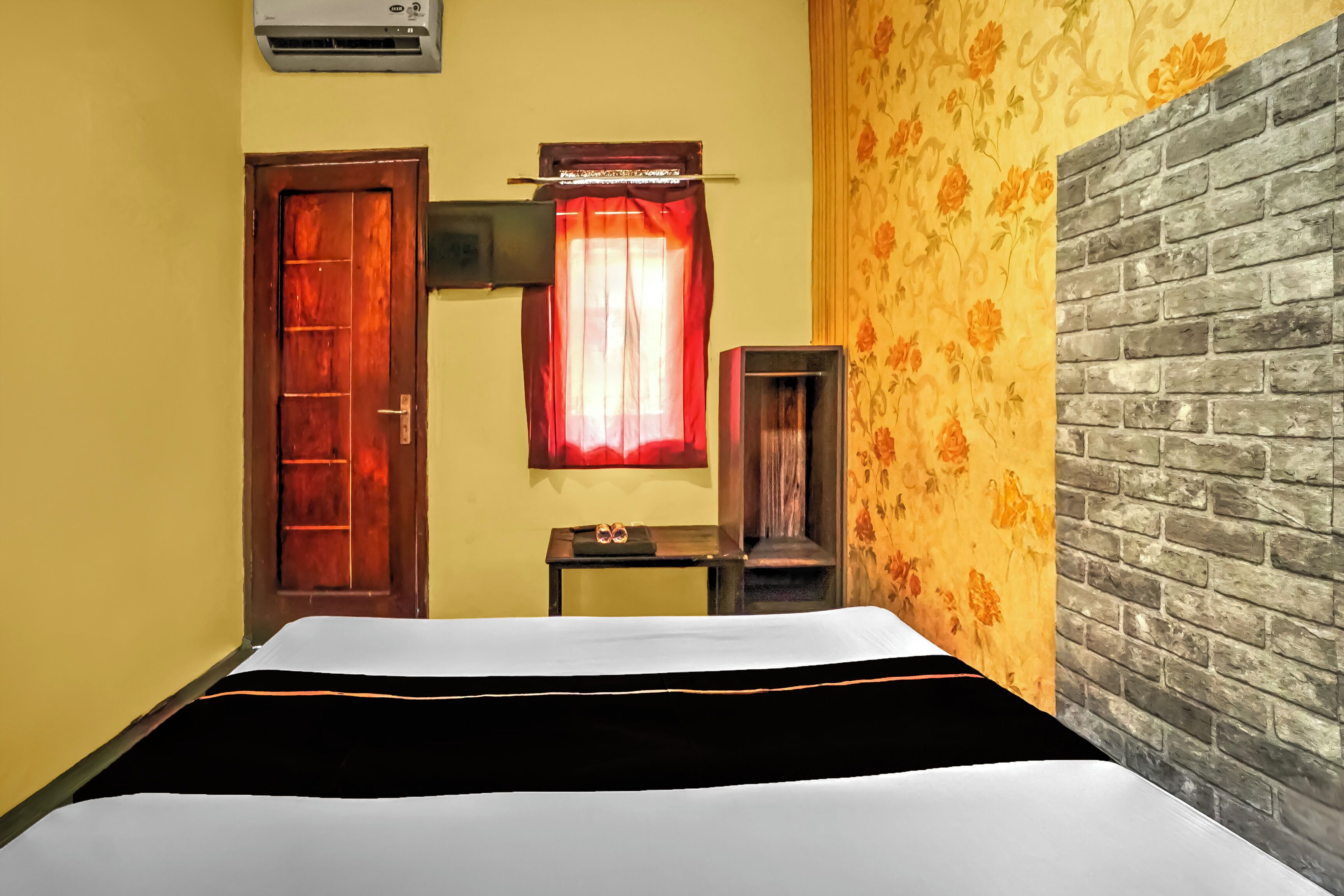 A hotel room with a bed, a wooden door, a window with a red curtain, a small table with a mirror, and a brick-patterned wall.