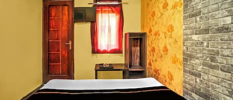A hotel room with a bed, a wooden door, a window with a red curtain, a small table with a mirror, and a brick-patterned wall.