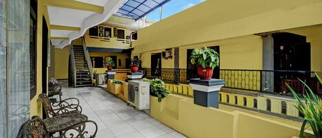 A spacious indoor courtyard with yellow walls, a tiled floor, and a glass ceiling. There are potted plants and a staircase leading to an upper level.