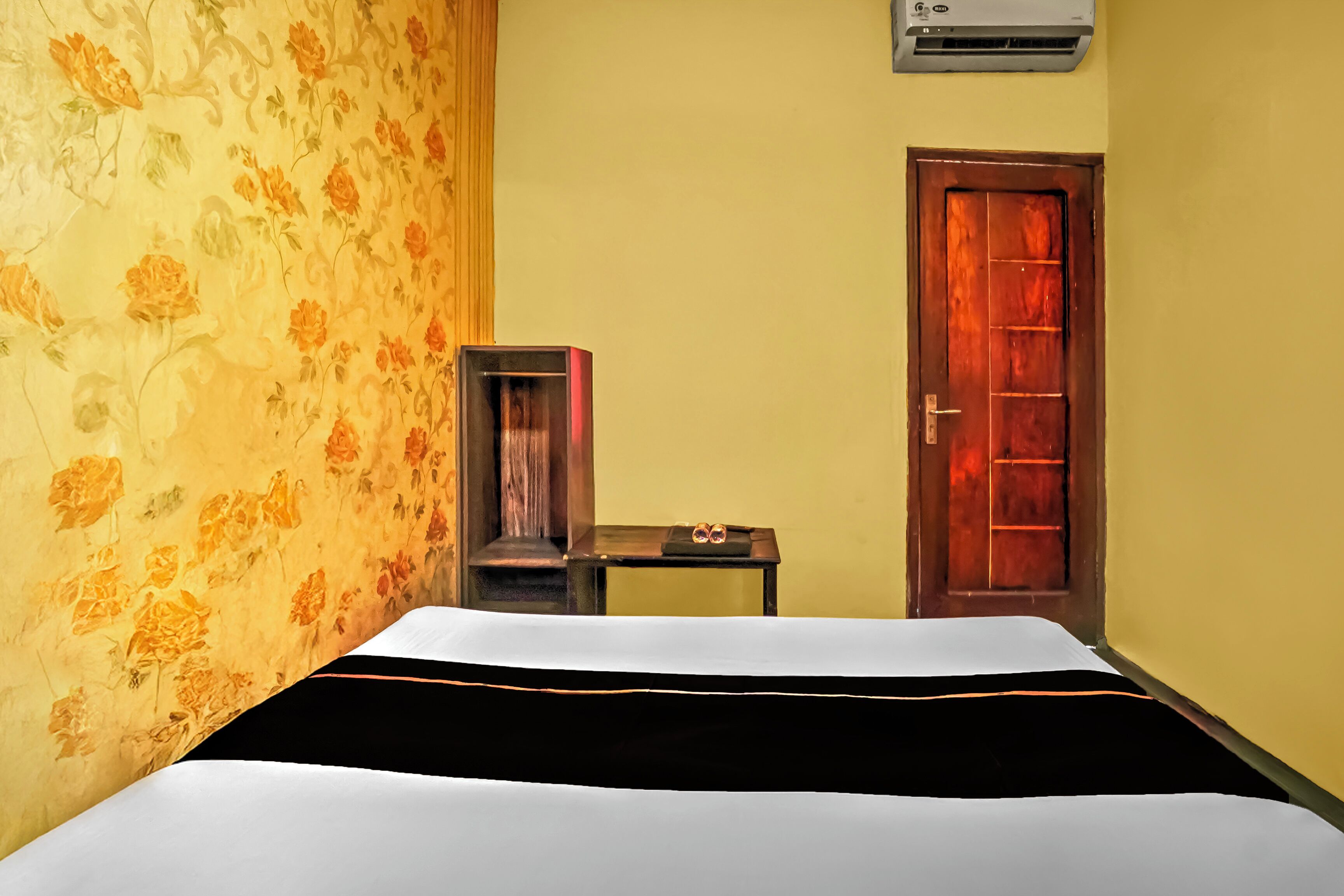 Quarto de hotel com uma cama, uma pequena mesa com um par de óculos, uma porta de madeira e um aparelho de ar condicionado na parede.