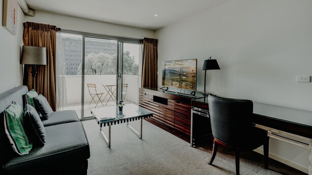 Ett modernt vardagsrum med en soffa, ett soffbord i glas, ett skrivbord och en stol. Det finns ett stort fönster med gardiner, en tavla på väggen och utsikt över byggnader utanför.
