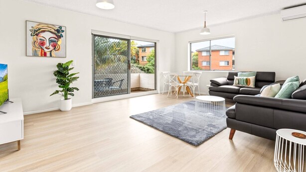 A modern living room with a sofa, a coffee table, and a dining area with chairs. There is a large window providing a view of the outside. - Bright and convenient apartment, close to shops (Ryde)