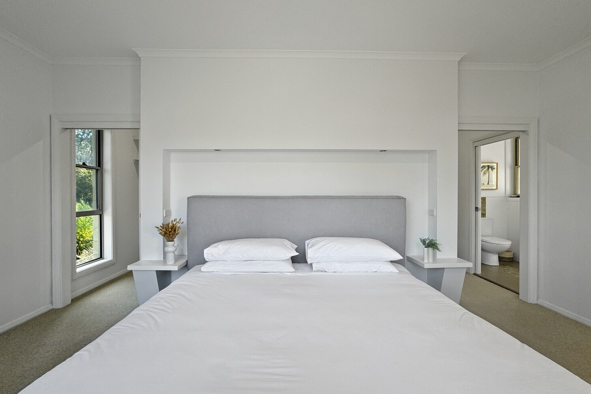A modern bedroom with a large bed, white walls, and a view of the outdoors through a window.
