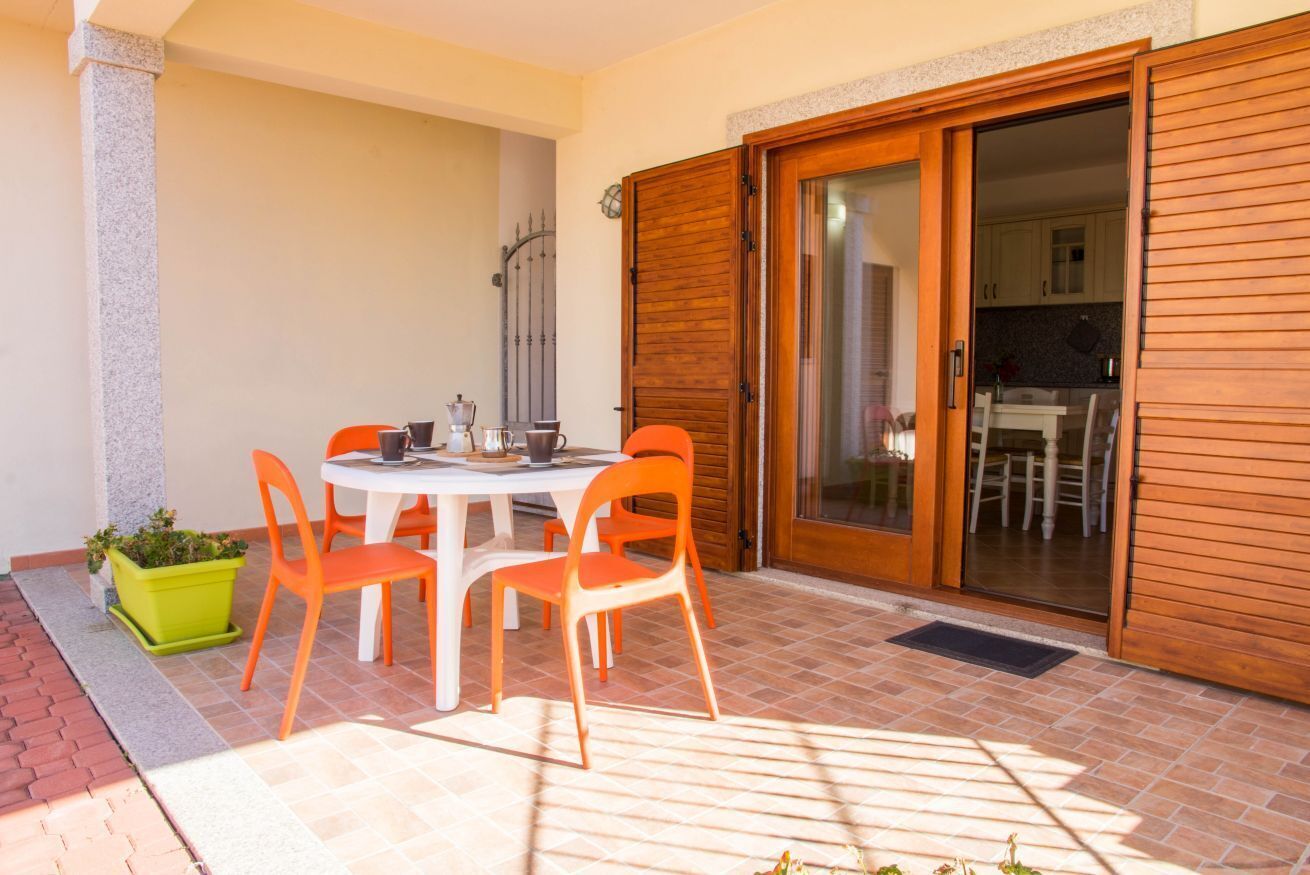 Um pátio externo com uma mesa redonda e cadeiras laranjas, um cachepot verde e uma porta de vidro deslizante que dá para uma cozinha.