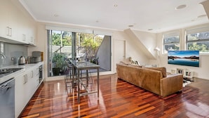 A modern kitchen with a dining area, a sofa, and a TV in a spacious living room with wooden flooring and large windows. - Stunning 2-Bed Unit with BBQ Patio near Dining (Northcote)