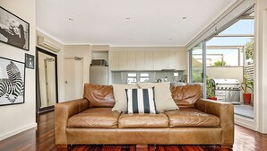 A spacious living room with a leather sofa, a large window, and a view of the backyard. - Stunning 2-Bed Unit with BBQ Patio near Dining (Northcote)