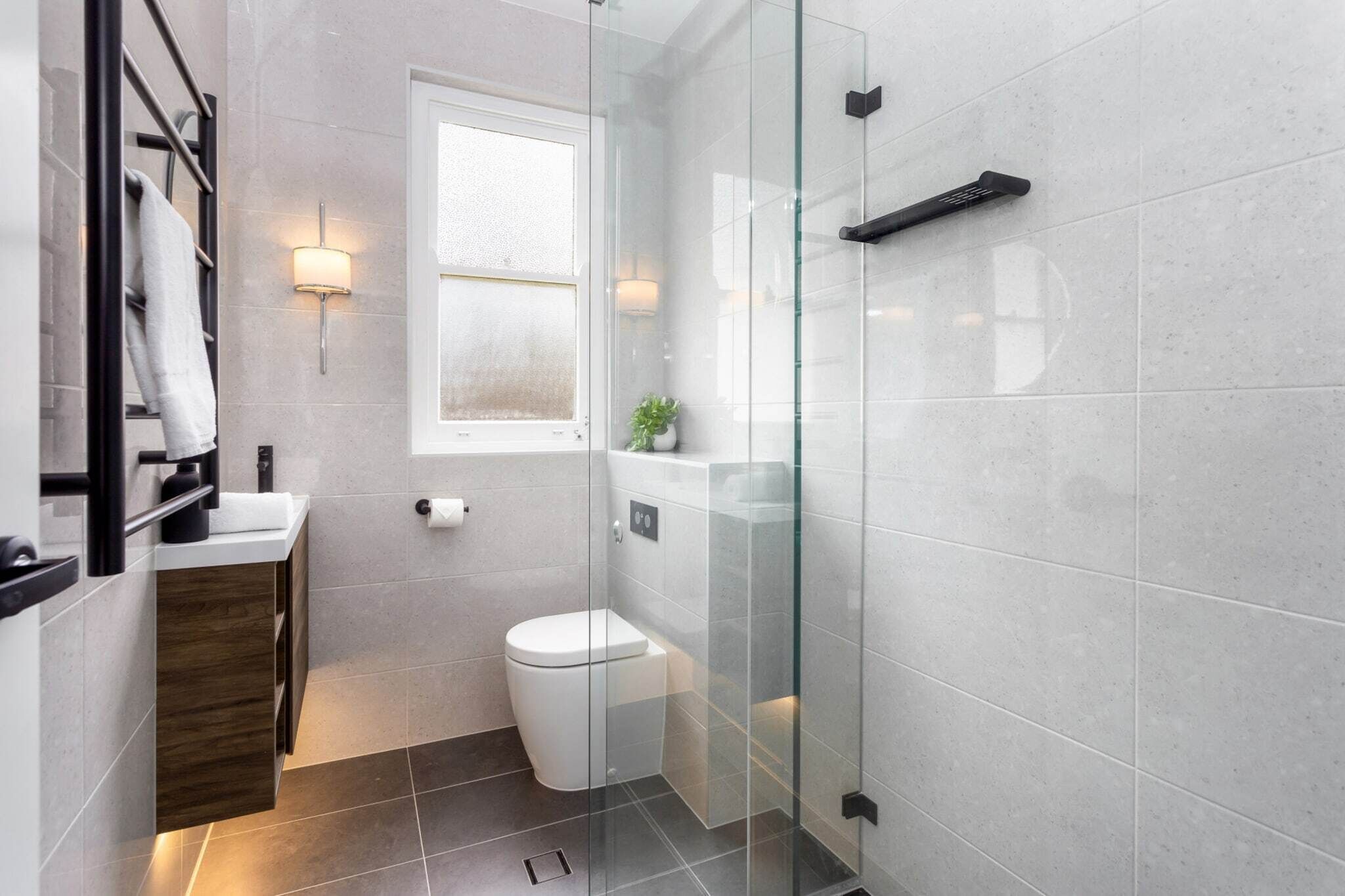 Une salle de bain moderne avec une douche à l’italienne en verre, des toilettes blanches et une petite fenêtre.