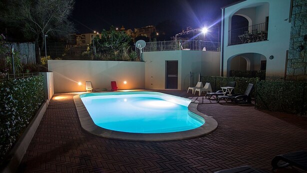 A swimming pool with a brick patio, lounge chairs, and a two-story building in the background. - GF Apartments La Vigna - Vigna Superior - LEVANTE (BADESI)
