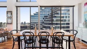 Uma sala de jantar moderna com uma mesa de madeira e cadeiras pretas, grandes janelas com vista para a cidade, e decoração minimalista com um quadro e um vaso com velas.