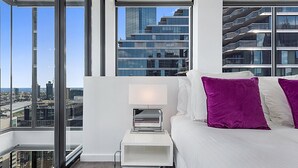 Quarto de hotel moderno com vista para a cidade, cama branca com almofadas roxas, mesa de cabeceira pequena com abajur e uma janela grande.