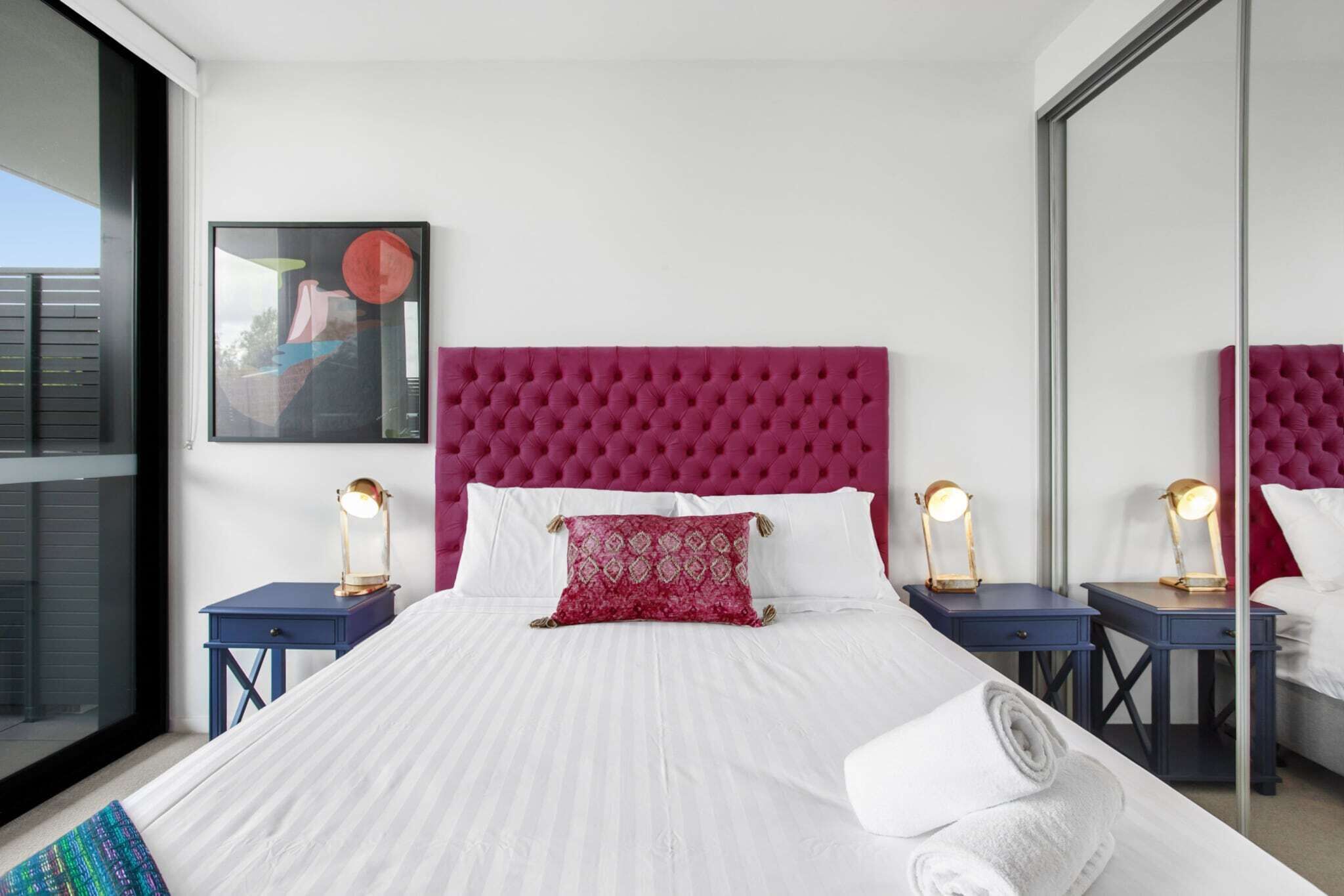 A modern hotel room with a large bed, white linens, a red tufted headboard, and two bedside tables with lamps.