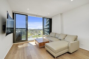 A modern living room with a large window, a beige sofa, a wooden coffee table, and a flat-screen TV mounted on the wall. - Modern 1-Bed Apartment With Parking (Phillip)