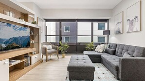 A modern living room with a large flat-screen TV, grey sectional sofa, wooden shelving, and a large window with a view of buildings. - Executive Unit with Pool and Gym near Riverfront (Southbank)