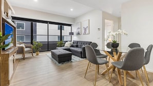 A modern living room with a sectional sofa, a glass dining table, and grey chairs. There is a TV on a wooden stand, a plant, and a large window with blinds. - Executive Unit with Pool and Gym near Riverfront (Southbank)