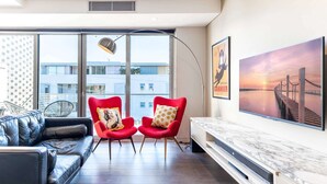 Una sala de estar moderna con un sofá de cuero negro, dos sillones rojos con cojines estampados, un ventanal con vistas a edificios, una lámpara de pie y un cuadro grande enmarcado de un atardecer sobre un muelle.