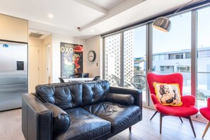 A modern living room with a black leather sofa, a red chair, and a large window offering a view of buildings. - Comfy 1-Bed Apartment in Perfect Location (Cremorne)