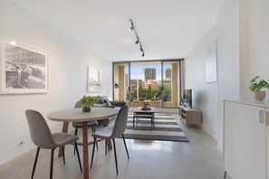 A modern living room with a dining area, large windows with a city view, a flat-screen TV, and a potted plant. - Central 1-Bedroom Apartment with City Views (Surry Hills)