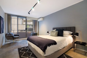 A modern bedroom with a large bed, a chair, a nightstand, and a window with a view. - Central 1-Bedroom Apartment with City Views (Surry Hills)