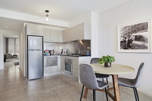 A modern kitchen with stainless steel appliances, a dining area with a round table and chairs, and a framed photo on the wall. - Central 1-Bedroom Apartment with City Views (Surry Hills)