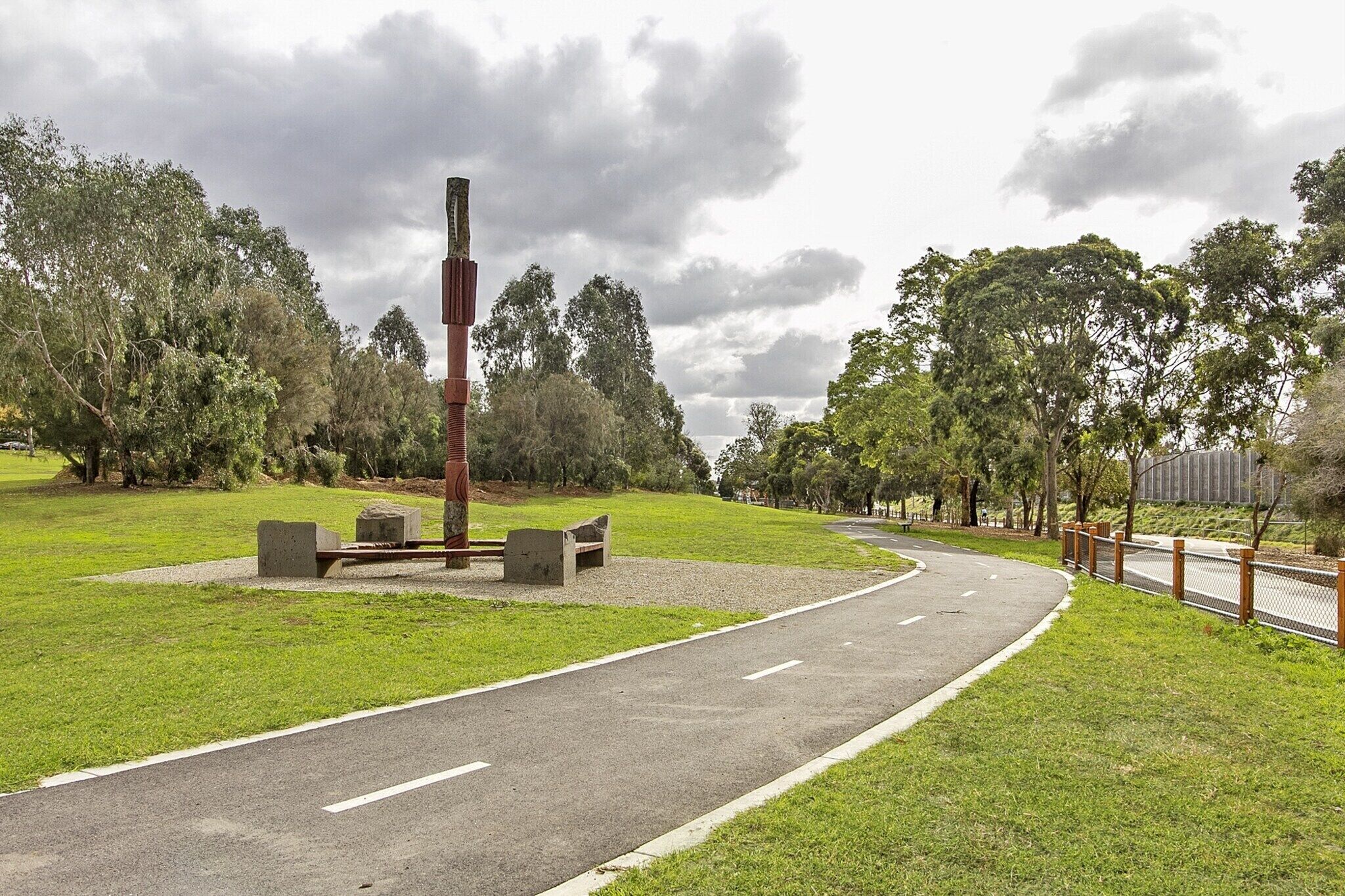 Un parc amb un camí sinuós, bancs i una escultura alta.