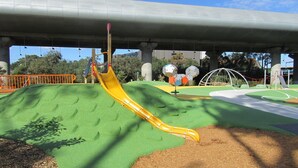 Ein Spielplatz mit einer gelben Rutsche, grünen Fallschutzbelägen und einer Kletterkonstruktion mit Netz.