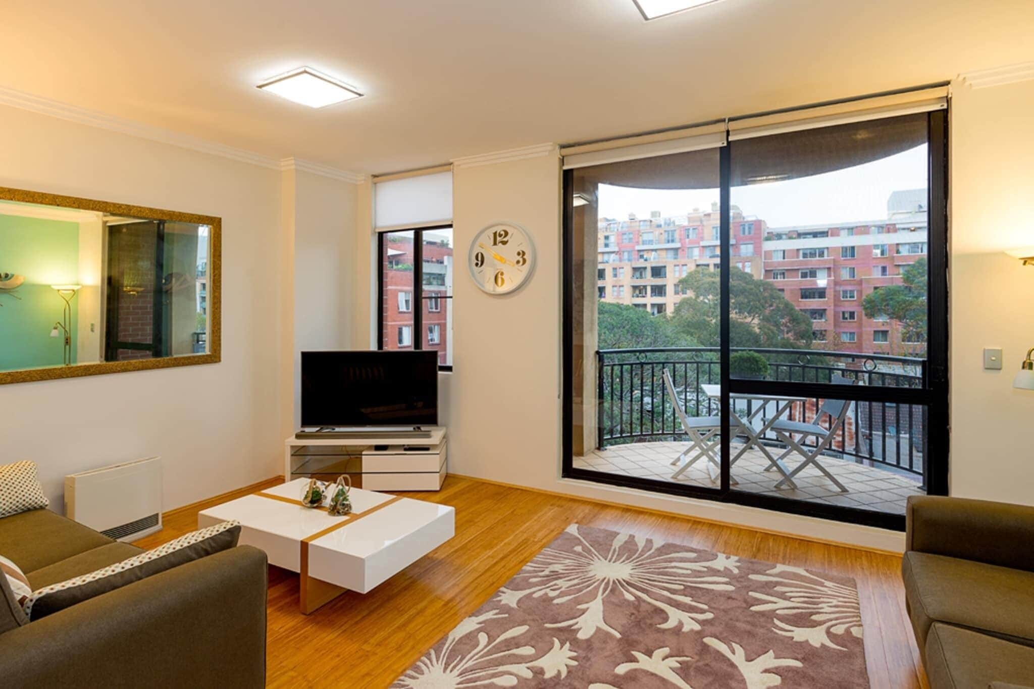 A modern living room with a flat-screen TV, a large window with a view of buildings, a clock, and a patterned rug.