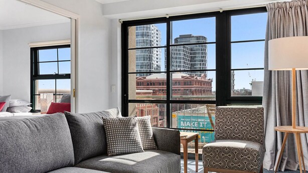 Una habitación de hotel moderna con un sofá gris, un sillón estampado y vistas a edificios de la ciudad a través de amplias ventanas.