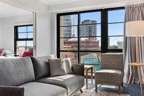 Una habitación de hotel moderna con un sofá gris, un sillón estampado y vistas a edificios de la ciudad a través de amplias ventanas.