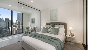 Een moderne slaapkamer met een groot bed, stadszicht door raamhoog-tot-vloer glas, een stoel en een klein nachtkastje met een lamp.