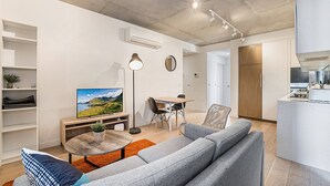 Ein modernes Wohnzimmer mit grauem Sofa, einem Couchtisch und einem Fernseher. Es gibt einen Essbereich mit Tisch und Stühlen sowie eine Küche mit weißen Schränken und einem Herd.
