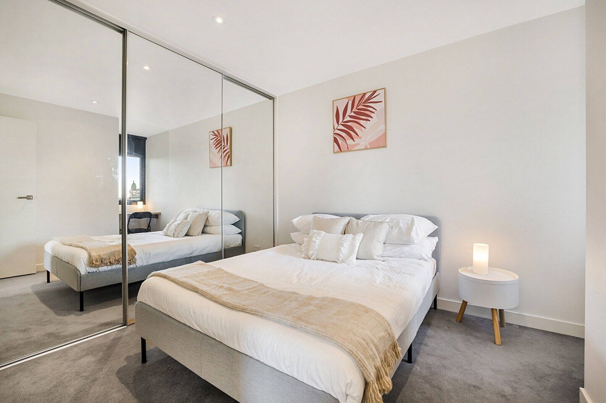 A modern bedroom with a large mirror, two beds, a bedside table with a lamp, and artwork on the wall.