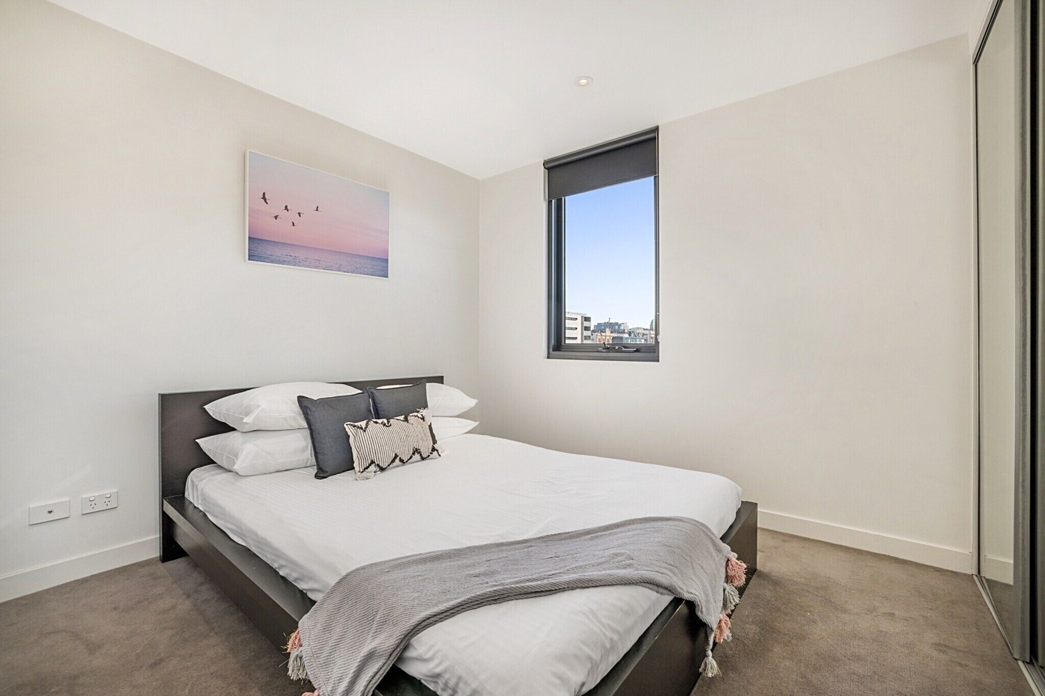 A modern bedroom with a bed, a city view through a window, and a painting on the wall.