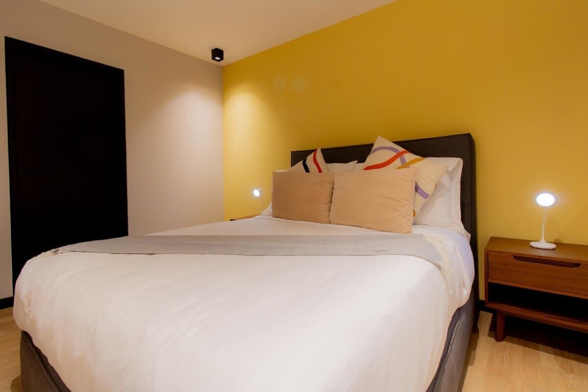 Un letto rifatto con cura, lenzuola bianche e numerosi cuscini, un comodino con una lampada e una porta scura.