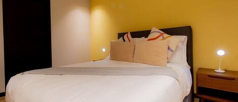 Una cama bien hecha con ropa blanca y varios almohadones, una mesita de noche con una lámpara y una puerta oscura.
