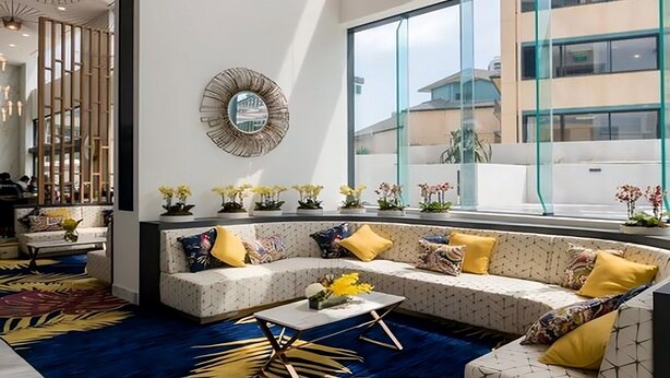 En modern hotellobby med ett stort fönster, en dekorativ spegel och en sittgrupp med kuddar och blommor.