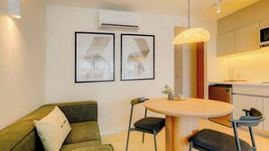 A modern living room with a green sofa, a round wooden table, and two chairs. There are two framed pictures on the wall, an air conditioning unit, and a kitchen area with a microwave. - TOP The Raven Double | Modern 2BR apartment (Mexico City)
