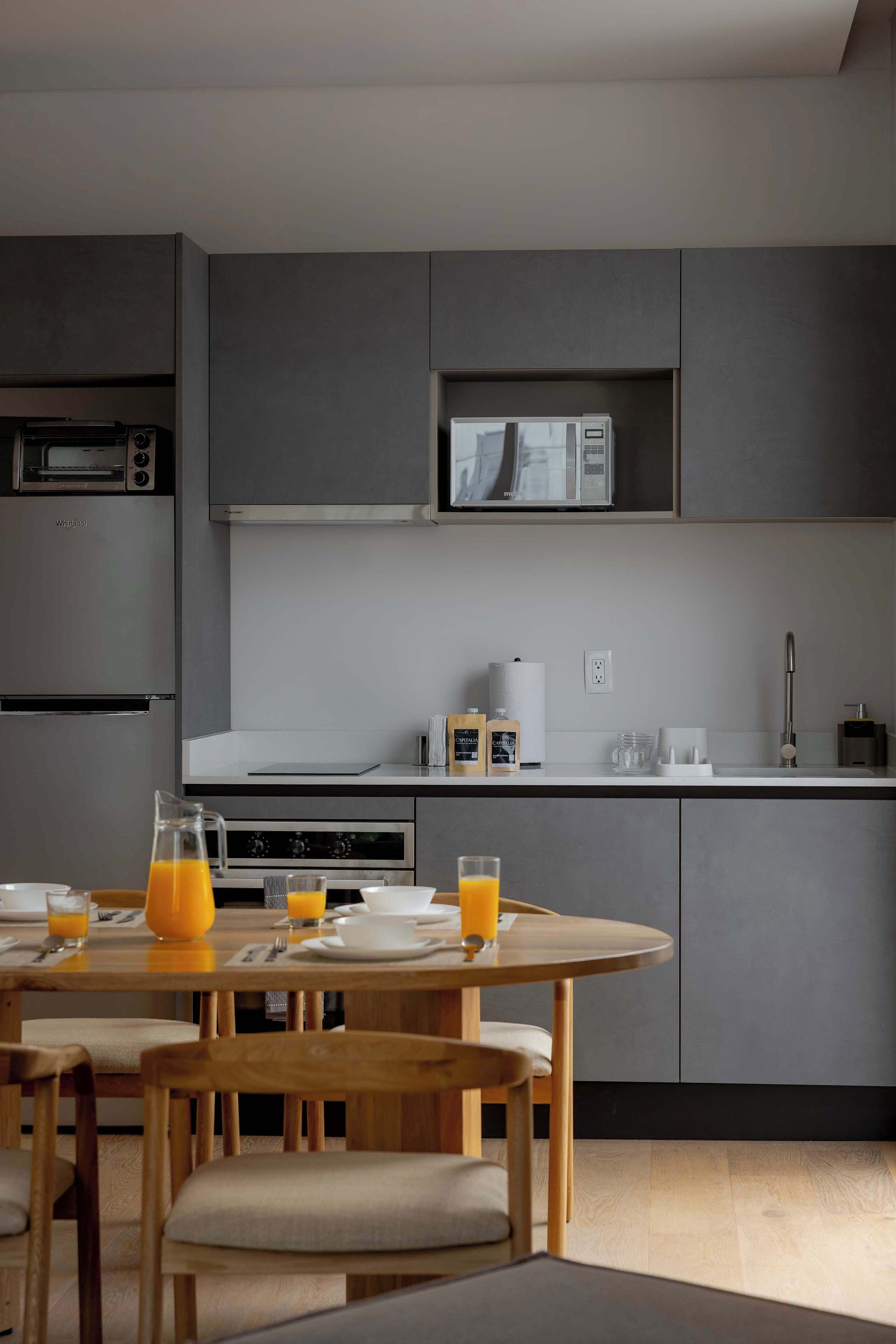 A modern kitchen with grey cabinets, a microwave, and a coffee maker. There is a dining table with chairs and a pitcher of orange juice.